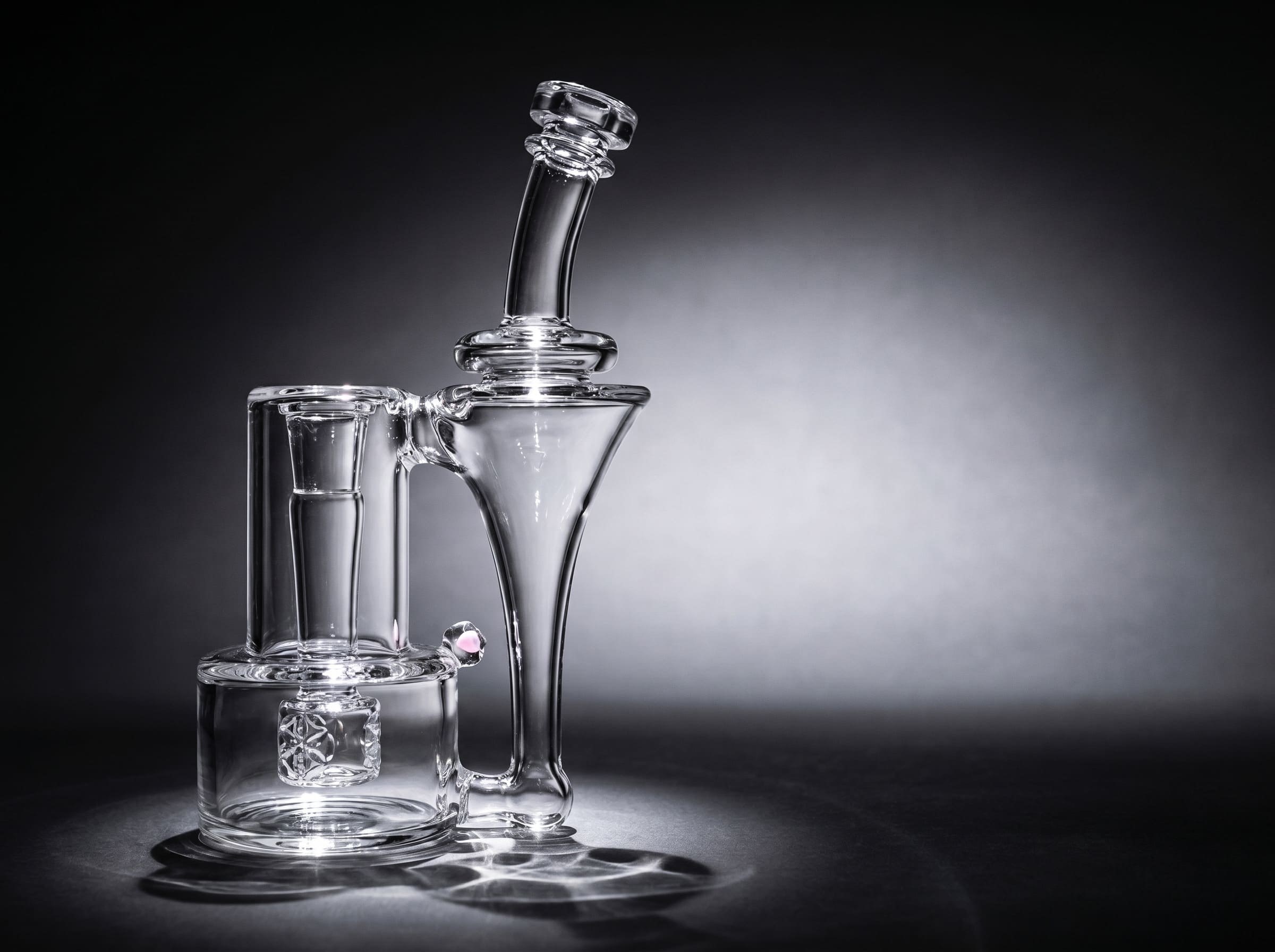 A clear glass recycler dab rig lit by a spotlight on a black background
